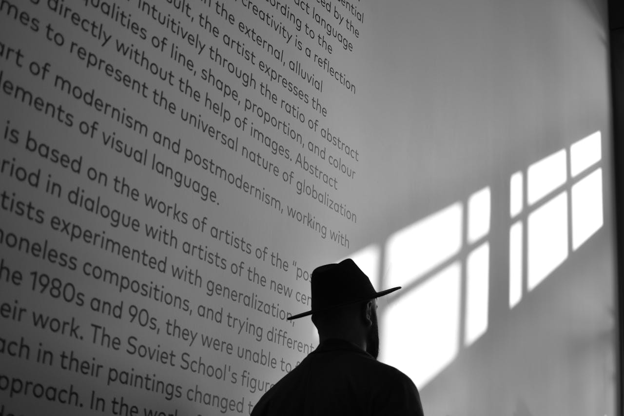 A person in hat observes museum wall with text and light shadow.
